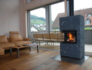 Dunkler, moderner Specksteinofen. Es brenn ein Feuer darin. Er steht vor einer grossen Fensterfront mit Balkon. Links im Bild steht eine beige Lederpolstergruppe. Auf dem Balkon steht ein Holzbank und man sieht aufs Nachbarhaus und links auf Landschaft